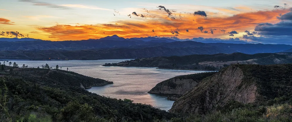 Lake winding between dark chaparral ridges under an orange sunset sky